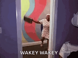 a woman is standing in a doorway with a broom in her hand and the words wakey wakey on the wall behind her .