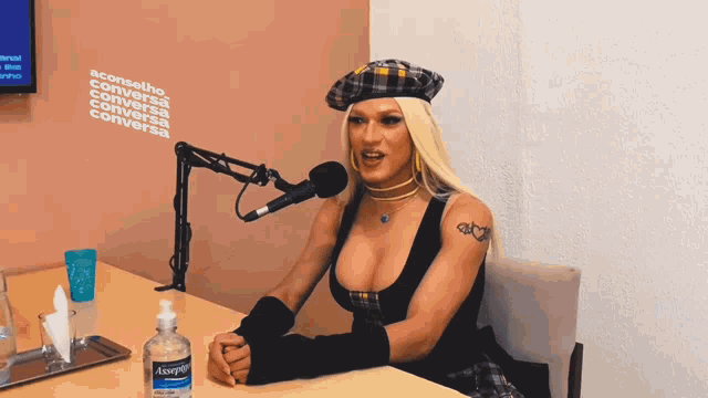 a woman in a plaid hat sits at a table with a bottle of hand sanitizer in front of a microphone