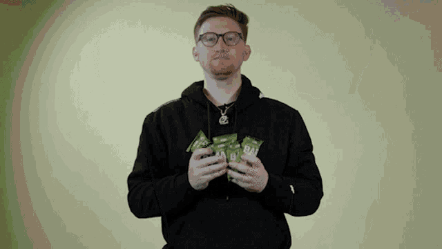a man wearing glasses and a black hoodie holds up a few packets of bacon