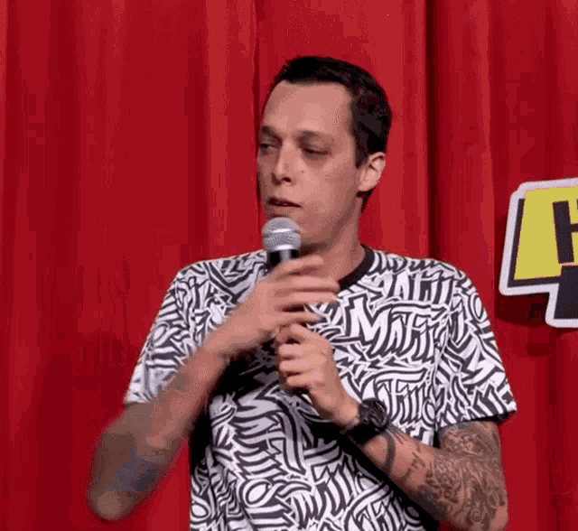 a man in a black and white shirt with the letter m on it holds a microphone in front of a red curtain