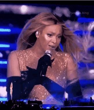 a woman is singing into a microphone on a stage while wearing a gold dress and black gloves .