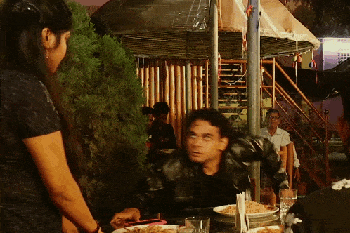 a man sits at a table with plates of food and a woman standing behind him