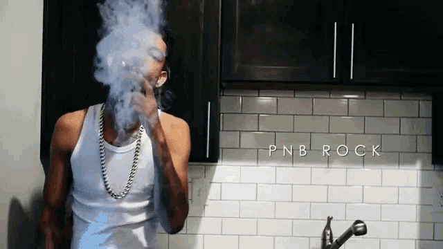 a man in a white tank top is smoking a cigarette in a kitchen with pnb rock written on the wall