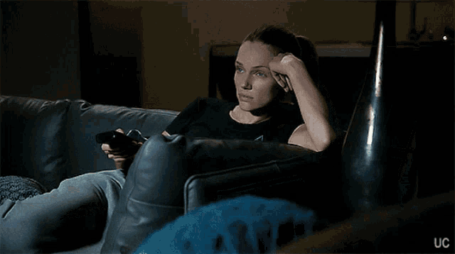 a woman sitting on a couch holding a remote control with uc written on the bottom right