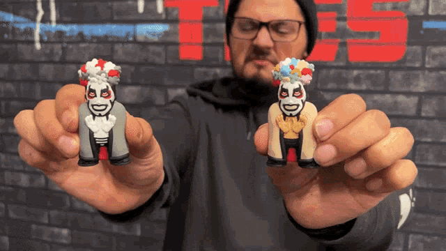 a man holds two toy figures in front of a brick wall with the letter ts on it