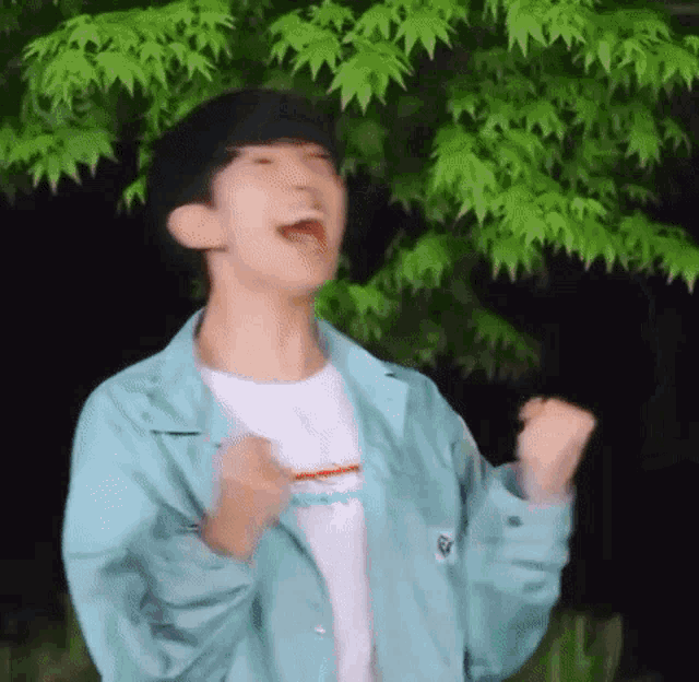 a young man in a blue jacket is standing in front of a tree with his hands in the air .