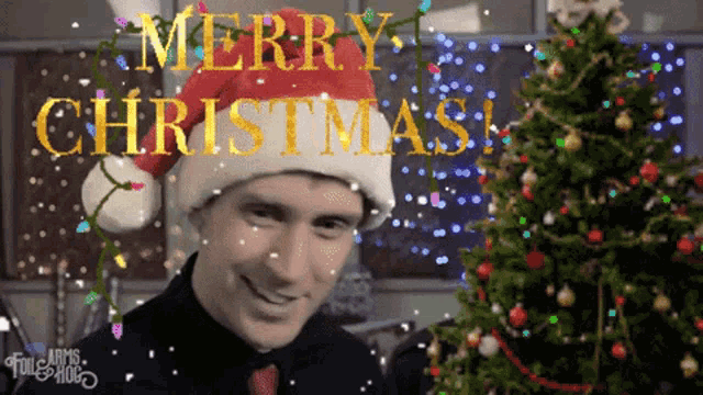 a man wearing a santa hat is standing in front of a christmas tree with merry christmas written on it