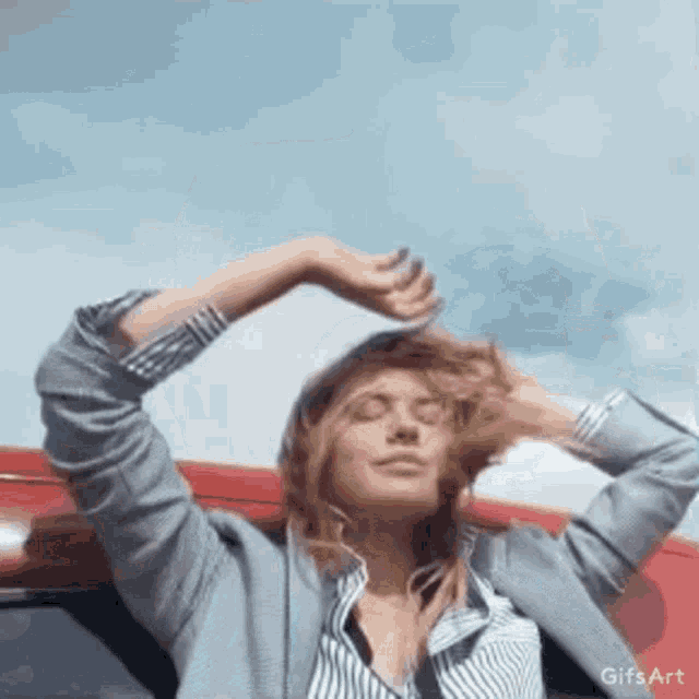 a woman is standing in front of a red car with her eyes closed and her hair blowing in the wind .
