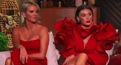 two women in red dresses are sitting on a couch and looking at something .
