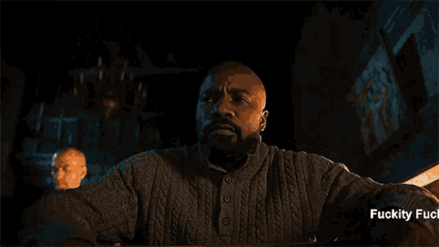 a man with a beard is sitting at a table in a dark room with the words fuckity fuck fuck fuck .