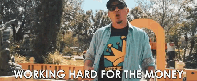 a man wearing sunglasses and a t-shirt that says outk working hard for the money stands in a park