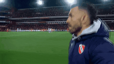 a man with a beard wearing a blue jacket with a red shield on it stands on a soccer field