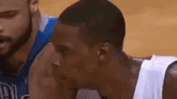 a man in a white tank top is sitting on a basketball court next to another man in a blue jersey .