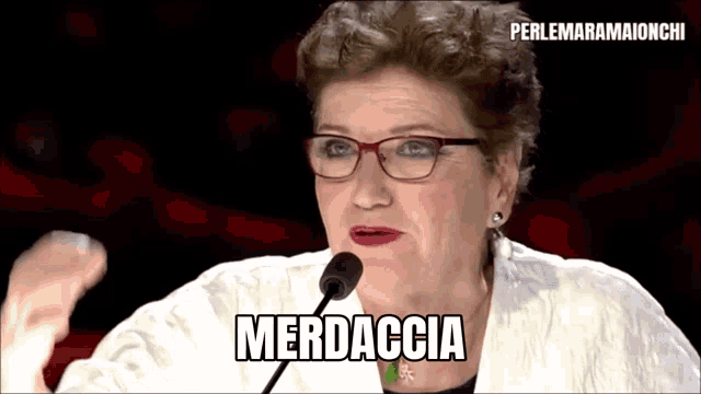 a woman wearing glasses is giving a speech in front of a microphone and saying merdaccia .