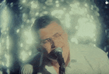 a man with glasses singing into a microphone with the letter i on the bottom right