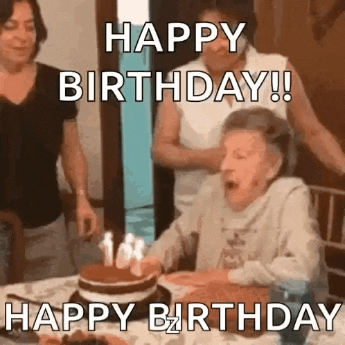 an elderly woman is blowing out candles on a birthday cake while sitting at a table .
