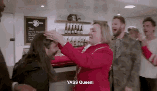 a woman in a red jacket is putting a crown on a man 's head and says yass queen