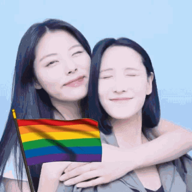two women are hugging each other with a rainbow flag in the background .