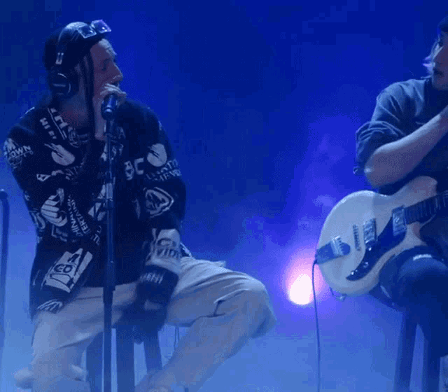 a man singing into a microphone while another man plays a guitar with a sticker that says ' us '