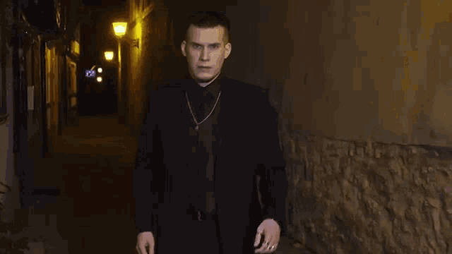 a man in a black suit and tie is standing in a dark alleyway at night .