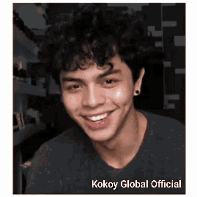 a young man with curly hair is smiling and wearing earrings and a black shirt .