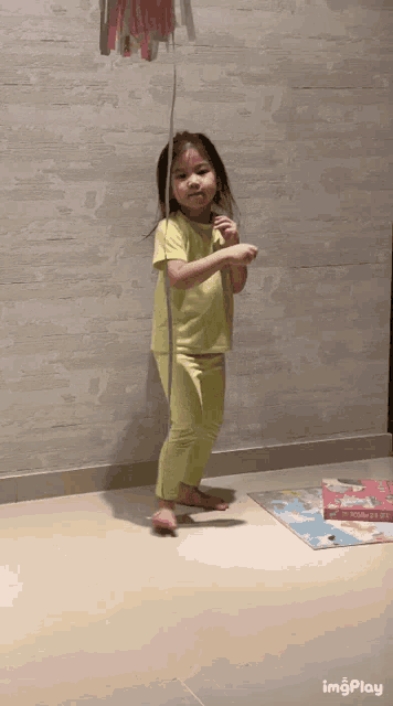 a little girl in a yellow shirt is standing on a tiled floor holding a string