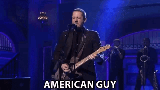 a man in a suit is playing a guitar and singing into a microphone with the words american guy below him .