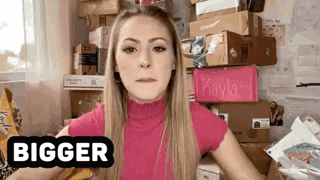 a woman in a pink shirt is standing in front of a pile of boxes and a sign that says " bigger "