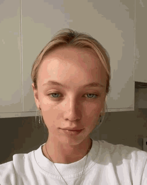 a young woman with blonde hair and blue eyes is wearing a white shirt and necklace .