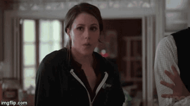 a woman in a black hoodie is standing in a room with a man in a plaid shirt behind her .
