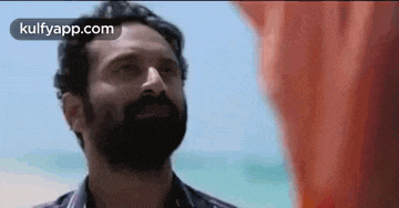 a man with a beard is standing on a beach with a red cloth in the background .