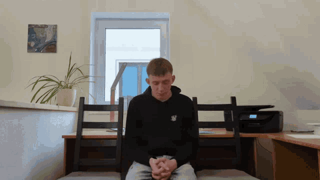 a man in a black sweatshirt with the letter s on it sits in front of a window
