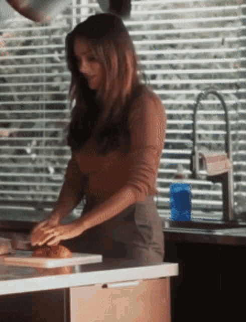 a woman is standing in front of a window with blinds while preparing food