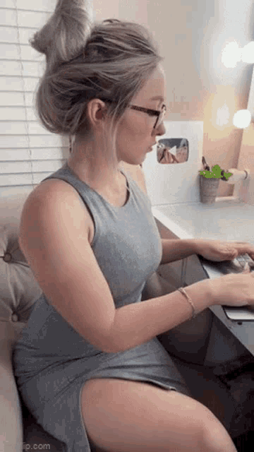 a woman in a gray dress and glasses is sitting at a desk using a laptop computer .