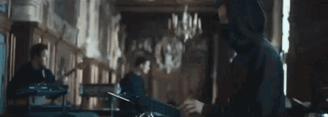 a man in a hooded jacket is playing a piano in a room with other people .