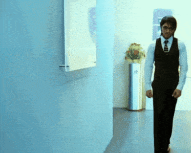a man in a vest and tie walks down a hallway with a hp trash can in the background