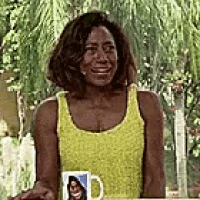 a woman in a yellow tank top is smiling while sitting at a table with a mug .