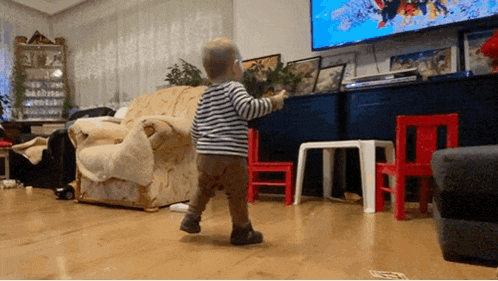 a baby is walking in a living room in front of a large flat screen tv