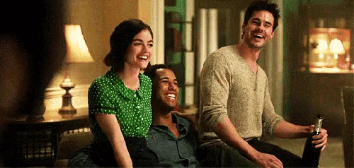 three people are sitting on a couch laughing and one of them is holding a bottle of beer