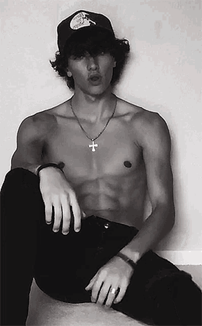 a shirtless man wearing a hat and a cross necklace is sitting on a bed .