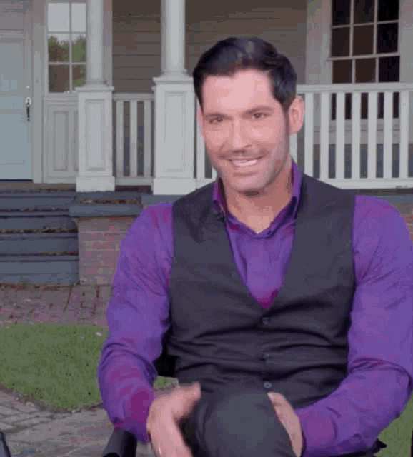 a man in a purple shirt and black vest smiles while sitting in front of a house