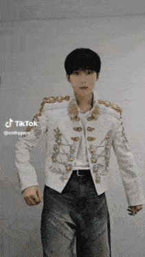a young man wearing a white jacket and jeans is standing in front of a white wall .