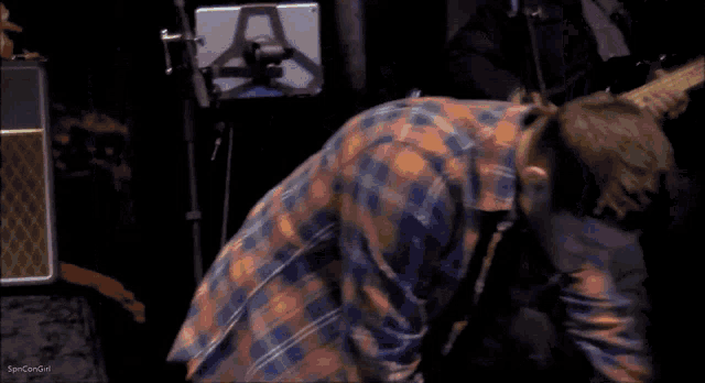 a man in a plaid shirt is laying on the floor while a man plays a guitar in the background