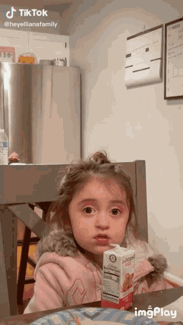 a little girl sitting at a table next to a carton of juice