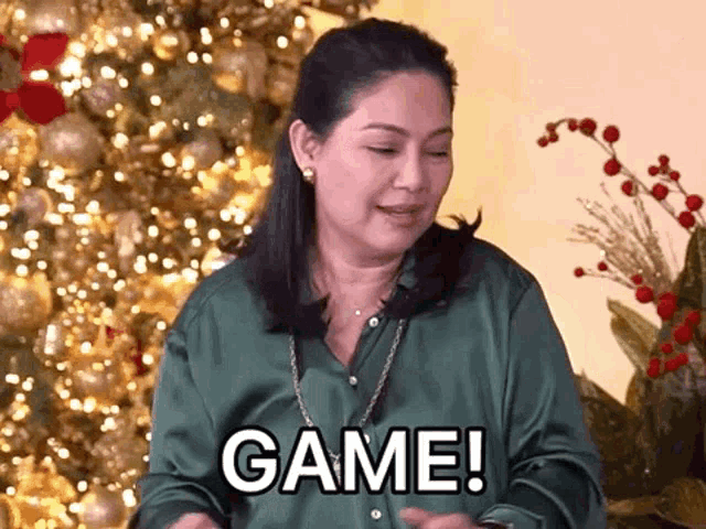 a woman in a green shirt is standing in front of a christmas tree and says game !