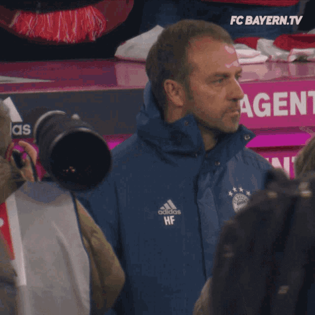 a man in a blue t-mobile jacket stands in front of a fc bayern.tv sign