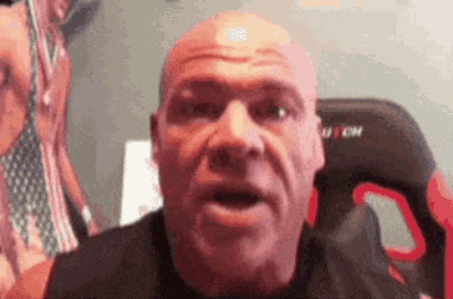 a bald man is making a funny face while sitting in front of a chair .
