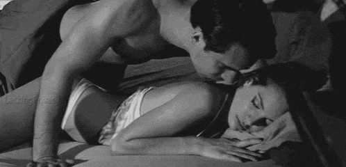 a black and white photo of a man kissing a woman on the neck while laying on a bed .