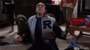 a man wearing a varsity jacket with the letter r on it is sitting on the floor .