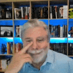 a man with a beard is touching his nose in front of a bookshelf filled with books .